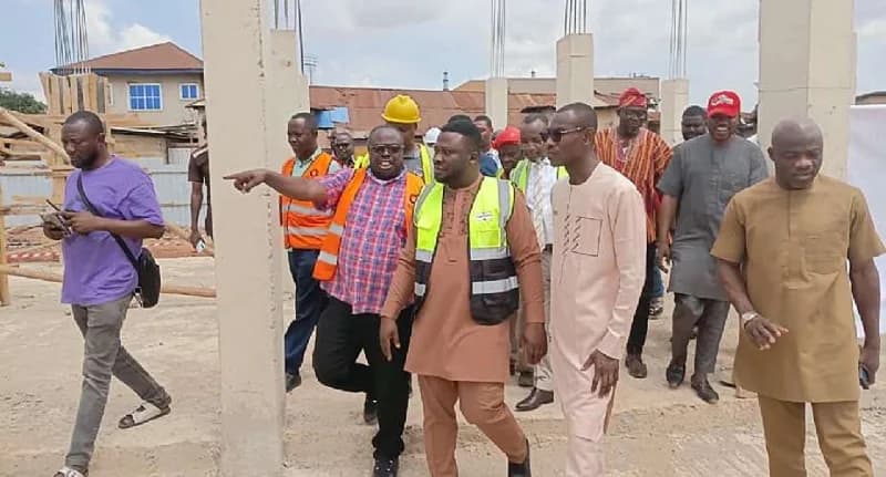 A group of officials and construction workers, including the Ashanti Regional Minister, inspect an ongoing market construction site. One man in an orange safety vest points toward the project's progress as they walk through the area featuring newly erected concrete pillars.