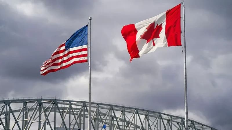 The flags of the United States and Canada fly side-by-side on tall flagpoles against an overcast sky. In the foreground, the top of a large steel truss bridge structure is visible, suggesting a border crossing or industrial area.
