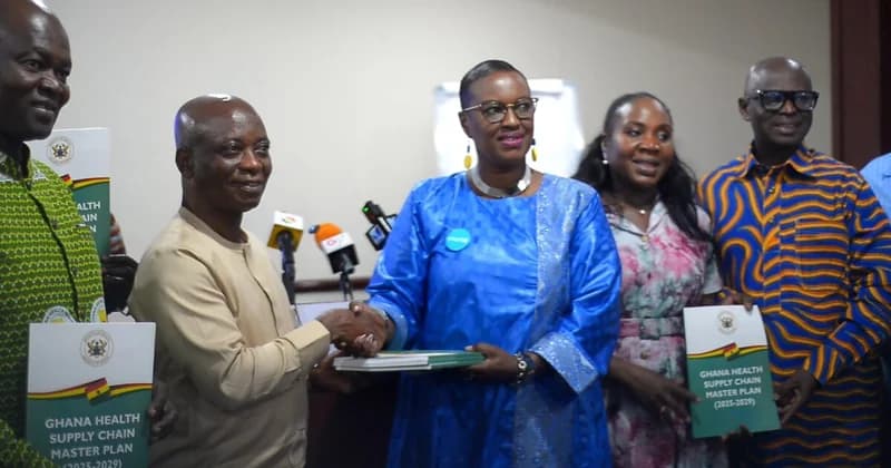 A group of Ghanaian officials and stakeholders are pictured during the official launch of the 'Ghana Health Supply Chain Master Plan (2025-2029)'. A man and woman in the center shake hands while holding the document, surrounded by others holding copies of the report in front of various news microphones.