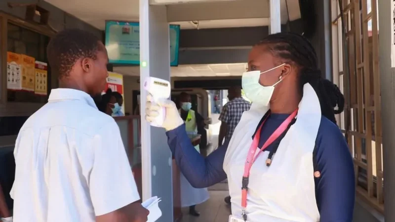 A health worker wearing a protective mask, gloves, and apron uses a handheld infrared thermometer to screen a man at a public entry point. In the background, posters with 'EBOLA' written on them are visible, alongside a metal detector frame and other health personnel.
