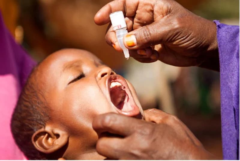 A close-up photograph shows a young child receiving oral polio vaccine drops from a healthcare worker. The image captures the moment a drop is administered into the child's open mouth, highlighting public health efforts in immunization.