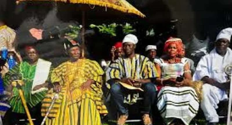 A group of traditional leaders and dignitaries are seated under a large yellow ceremonial umbrella at a public event. They are wearing colorful Ghanaian smocks and traditional attire, representing a cultural celebration likely associated with the Kakube festival.