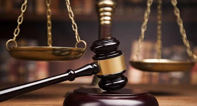 A close-up photograph of a dark wooden gavel with a gold band resting on a sound block. In the blurred background, the golden scales of justice are visible against a backdrop of what appears to be law books on a shelf.