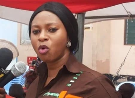 The image shows Sarah Adwoa Safo, the Member of Parliament for Dome-Kwabenya, speaking during a press engagement or public function. She is seen in a close-up shot with multiple microphones positioned in front of her, under a red and white tent structure.