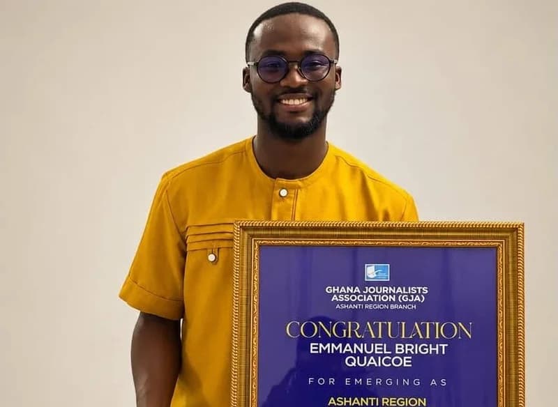 Emmanuel Bright Quaicoe, a journalist with Multimedia, is seen smiling while holding a framed award from the Ghana Journalists Association (GJA). He is wearing a mustard-yellow traditional shirt and glasses against a plain light-colored background.