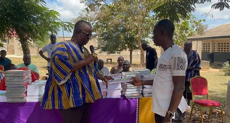 A man in a traditional blue and gold smock is presenting educational books to another person at an outdoor community event. Several stacks of books are visible on a table, and a small audience is gathered in the background under trees, suggesting an official donation or school ceremony.