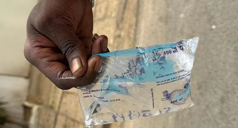 A close-up photograph of a person's hand holding a transparent 400ml plastic water sachet. The sachet features blue branding and French text, representing a common source of drinking water in West African nations like Guinea.