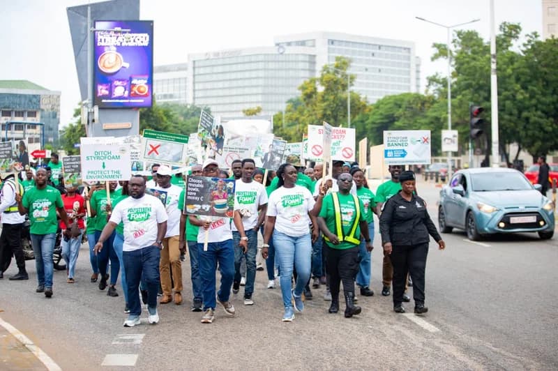 A group of people are seen marching through an urban street in Ghana, wearing green and white t-shirts with anti-styrofoam messages. They are holding several placards and banners that advocate for the ban of polystyrene foam, with a police officer walking alongside the group for security.