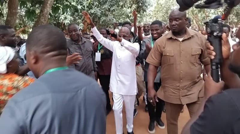 Johnson Asiedu Nketia, the National Chairman of the NDC, is captured walking through a crowd of supporters while gesturing with his arms raised. He is dressed in a signature white kaftan and is surrounded by security and party members wearing green lanyards, with a media camera visible in the foreground.