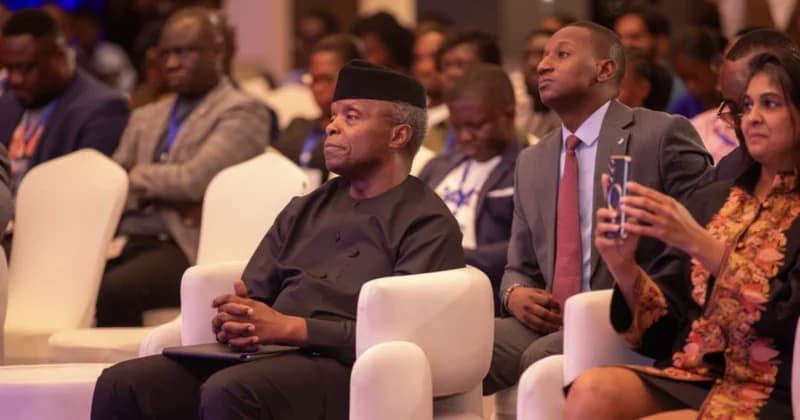 A photograph of attendees at a formal business conference or economic summit. The central figure, dressed in traditional attire with a black cap, sits attentively in the front row alongside other delegates in professional suits, while an attendee nearby uses a smartphone to capture the event.