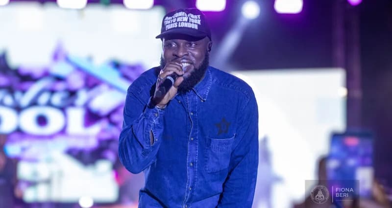 Ghanaian rapper M.anifest is seen performing live on stage during a concert celebrating the 10th anniversary of his 'Nowhere Cool' album. He is wearing a blue denim shirt and a black cap while rapping into a microphone against a backdrop of purple stage lights and a screen displaying the album's logo.