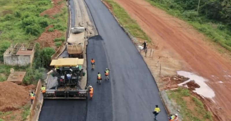 A high-angle photograph capturing active road construction on the Sunyani inner ring road. An asphalt paver is seen laying fresh black bitumen while a dump truck is positioned ahead of it, with several workers in high-visibility vests monitoring the progress.