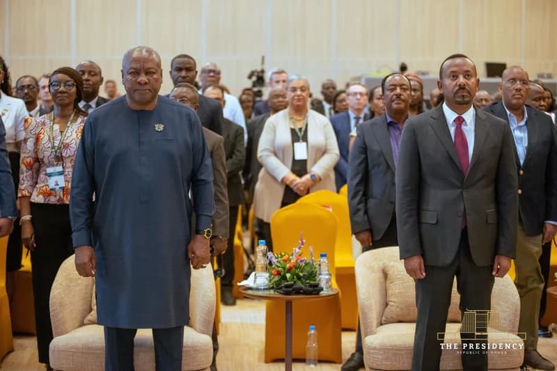 President John Mahama of Ghana and Prime Minister Abiy Ahmed of Ethiopia stand together during a formal ceremony at an international summit. They are positioned at the front of a hall filled with delegates and officials, signifying high-level diplomatic engagement within Africa.