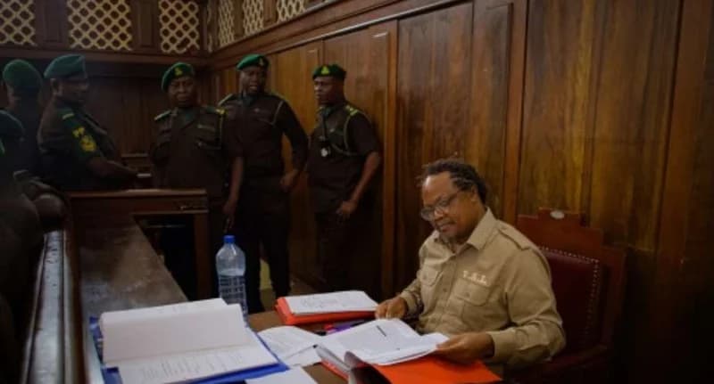 Tanzanian opposition leader Tundu Lissu is seen seated at a desk with several files and documents in a room with wood-paneled walls. Behind him, four uniformed security officers wearing green berets stand guard, suggesting a courtroom or custodial environment.