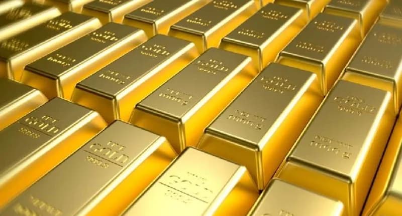 A close-up view of multiple rows of polished gold bars stacked together. Each bar features engraved text indicating its purity and weight, reflecting a bright yellow-gold metallic luster.