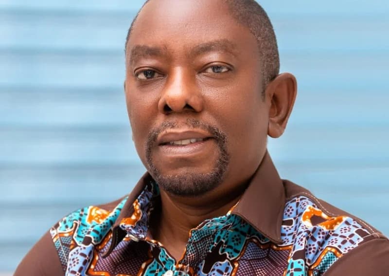 A close-up portrait of a man, likely the Ho Technical University lecturer mentioned in the article, looking towards the camera. He wears a brown shirt with intricate blue and orange patterns on the shoulders and collar against a soft blue background.