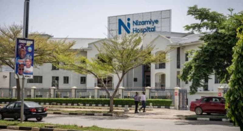 A wide-angle photograph of the Nizamiye Hospital in Abuja, Nigeria, featuring its white multi-story building and prominent blue signage. The foreground shows a paved street with a few passing vehicles and a streetlamp adorned with a political campaign poster for Bola Tinubu.