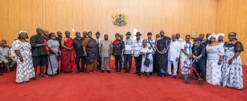 A large delegation of family members poses for a group photograph at Jubilee House following a formal meeting with government officials. Most individuals are dressed in traditional Ghanaian mourning attire of black, red, and white patterns, signifying the solemn purpose of their visit. The Ghana Coat of Arms is prominently displayed on the wooden paneled wall in the background above the group.