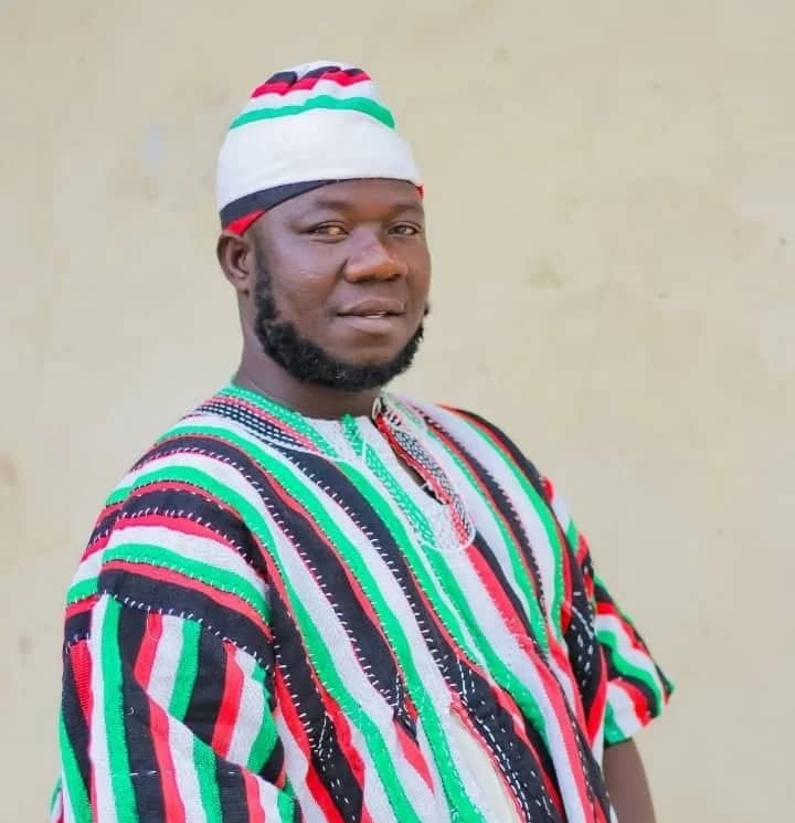 A portrait of Kwame Mborale Supply, the newly elected NDC chairman for Bimbilla. He is wearing a traditional Ghanaian smock (fugu) and matching hat featuring the red, green, white, and black colors of the National Democratic Congress (NDC).
