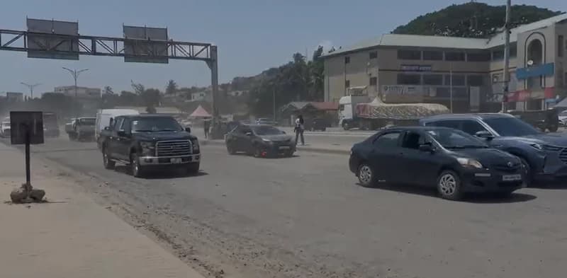 A view of a busy section of the McCarthy-Mallam road in Accra, showing vehicles navigating a dusty stretch of pavement. Several cars and a pickup truck are in motion, with commercial buildings and hills visible in the background under a bright sky.