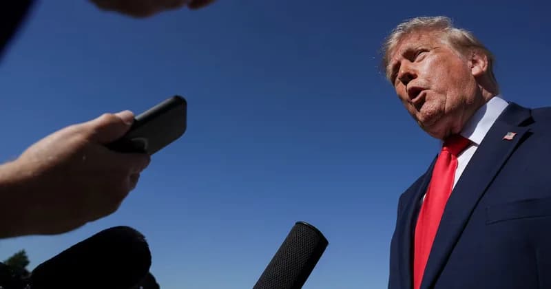 Donald Trump is pictured speaking outdoors against a clear blue sky. He is wearing a dark navy suit with a signature red tie and a small US flag pin on his lapel while addressing a group of reporters holding microphones and a smartphone.