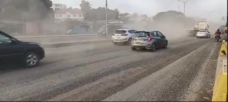 The image depicts vehicles navigating a dusty, deteriorating stretch of the McCarthy-Mallam road in Accra. A thick cloud of dust kicked up by traffic obscures the background buildings and trees, highlighting the poor infrastructure conditions that prompted public outcries.