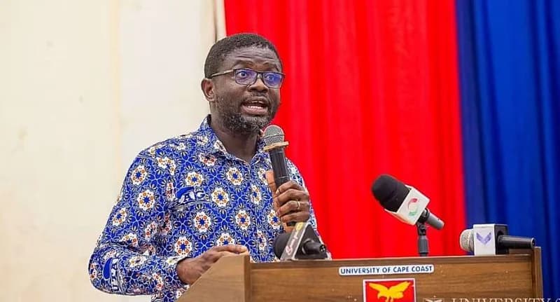 A man wearing glasses and a blue patterned shirt with the UTAG logo is seen speaking at a wooden podium labeled 'UNIVERSITY OF CAPE COAST'. He is holding a handheld microphone while several other media microphones are positioned on the lectern in front of him against a red and blue draped background.