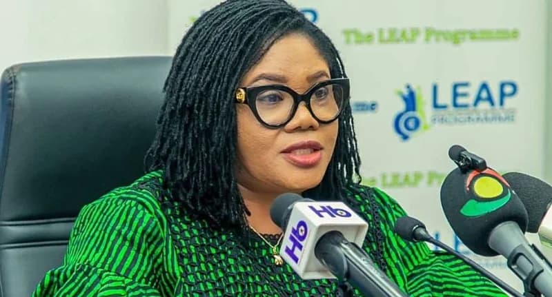 Dakoa Newman, Ghana's Minister for Gender, Children and Social Protection, is seen speaking into several microphones during a press event. She is wearing a green and black patterned dress and large black-framed glasses, seated in front of a banner for 'The LEAP Programme'.