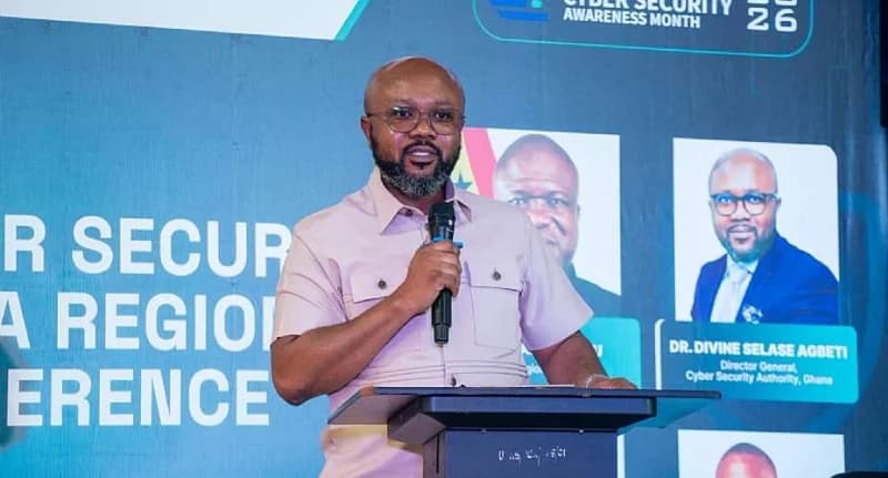 Dr. Divine Selase Agbeti, the Director-General of the Ghana Cyber Security Authority, is captured speaking at a podium during an official event. He is wearing a light pink button-down shirt and glasses, standing in front of a backdrop for a Cyber Security Awareness Month regional conference.