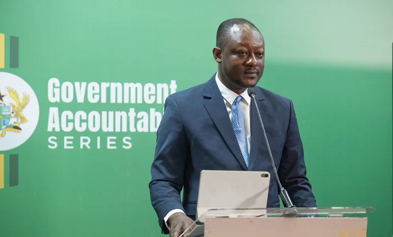Jerry Kofi Hinson, a former Managing Director of the Tema Oil Refinery (TOR), is seen speaking at a podium during a Government Accountability Series event. He is wearing a navy blue suit with a light blue patterned tie, standing in front of a green backdrop featuring the Ghana coat of arms and official event text.