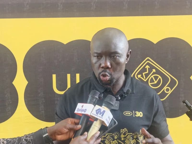 A man wearing a black MTN-branded polo shirt is captured speaking into several microphones during a media interview. He stands against a vibrant yellow and black MTN backdrop, featuring tech-related icons such as 'e-games' and 'AI'.
