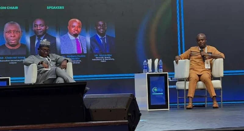 Dr. Mustapha Abdul-Hamid, Chief Executive of the National Petroleum Authority (NPA), is pictured speaking at the OTL Africa Downstream Energy Week. He is seated on a stage in a brown traditional outfit next to another participant, with a large screen in the background displaying speaker profiles and titles.