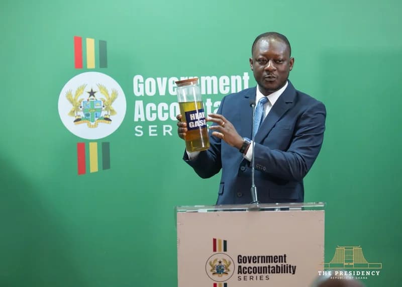 A man in a blue suit stands at a podium during a 'Government Accountability Series' event in Ghana. He is holding up a glass jar labeled 'HEAVY GAS OIL' containing a yellowish liquid, likely as a demonstration of refinery production. The background features the Ghanaian coat of arms and official government branding on a vibrant green backdrop.