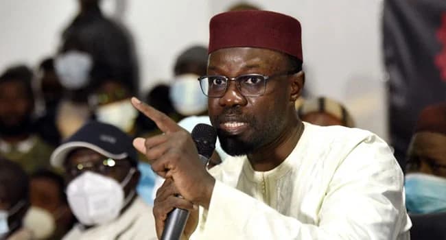 A close-up photograph shows Senegalese politician Ousmane Sonko speaking emphatically into a microphone while gesturing with his finger. He is wearing a traditional white boubou and a maroon kufi hat, with several people wearing face masks visible in the blurred background.