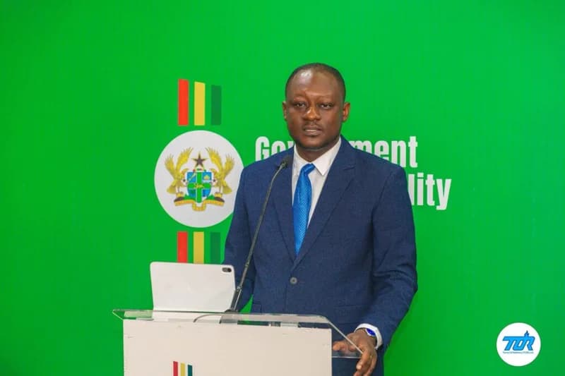 A man in a navy blue suit and bright blue tie stands behind a white podium while delivering a speech. The background is a solid green wall featuring the Ghana Coat of Arms and the words 'Government' and 'lity', with a Tema Oil Refinery (TOR) logo in the bottom right corner.