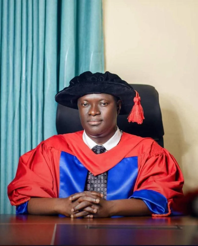 A formal portrait of Prof. Vincent Adzahlie-Mensah dressed in academic regalia, featuring a red and blue gown and a black academic cap with a red tassel. He is seated at a desk with his hands clasped, looking directly at the camera against a background of teal curtains.