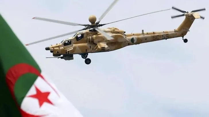 An Algerian military attack helicopter flies through a cloudy sky, with the national flag of Algeria partially visible in the blurred foreground. The aircraft features a desert camouflage pattern and displays the registration SC-55 on its tail boom along with the Algerian Air Force roundel.