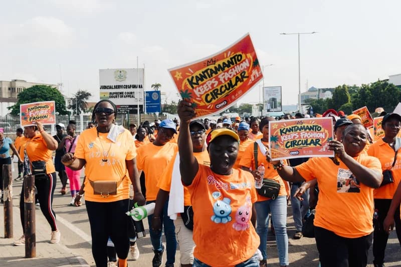 A large group of Kantamanto traders, mostly women wearing matching orange t-shirts, participate in a public march through the streets of Accra. Several participants hold up vibrant placards with slogans like 'Kantamanto Keeps Fashion Circular' as they pass the Judicial Service of Ghana building.