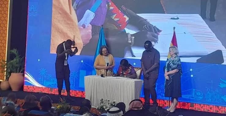 A high-level signing ceremony is captured on stage during a business summit in Ghana. A woman in traditional attire is seated at a table signing a document, while several international and local officials stand by as witnesses. A large LED screen in the background shows a live close-up of the signing process to the audience.