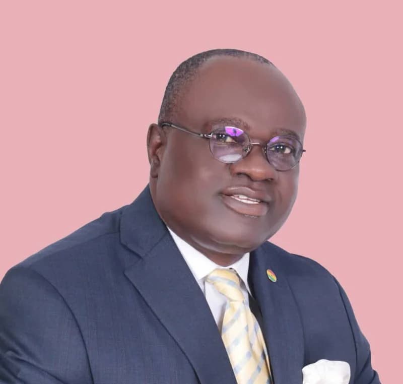 A formal studio portrait of Joseph Osei-Owusu, the First Deputy Speaker of the Parliament of Ghana. He is pictured wearing a dark blue suit, a yellow and blue striped tie, and spectacles against a solid pink background.