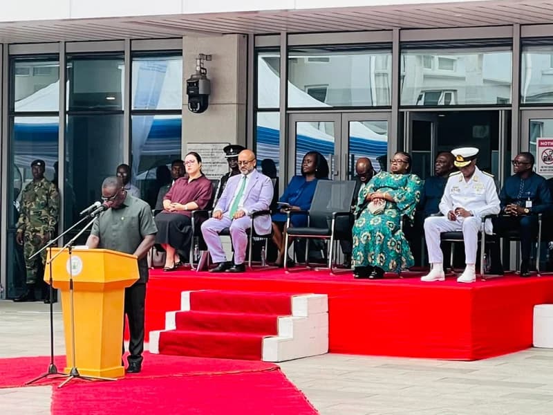 An official delivers a speech from a yellow podium during a formal government ceremony in Ghana. Behind the speaker, a group of dignitaries, including a high-ranking naval officer and individuals in traditional and formal Western attire, sit on an elevated stage covered in red carpet.