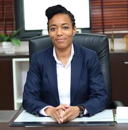 A professional portrait of Dr. Zanetor Agyeman-Rawlings, a Member of Parliament in Ghana, seated at an office desk. She is wearing a navy blue blazer and a white shirt with her hands clasped on the desk in a formal setting.