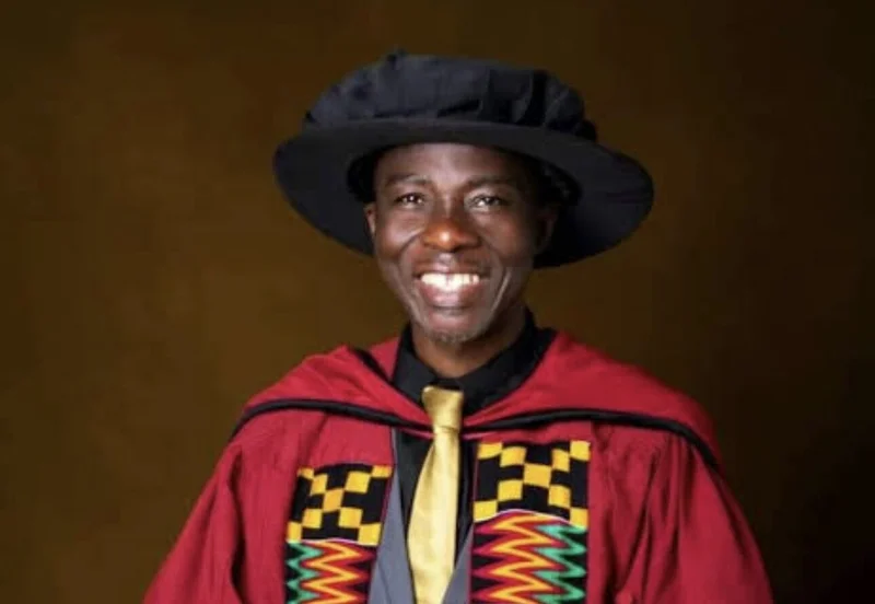 A professional studio portrait of Frederick Ofori-Nuako dressed in full academic regalia. He is wearing a red gown with distinctive kente-patterned lapels, a black shirt with a gold tie, and a black brimmed academic hat. The subject is smiling against a solid dark brown backdrop.