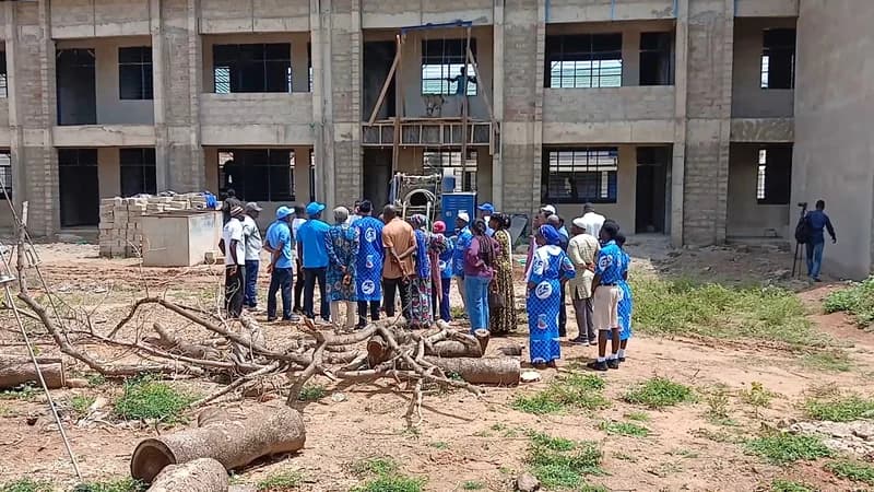 A large group of people, several wearing blue shirts featuring Ghanasco branding, are gathered at a construction site in front of an unfinished two-story concrete school building. The foreground is littered with cut logs and debris, illustrating the stalled state of the infrastructure project described in the article.