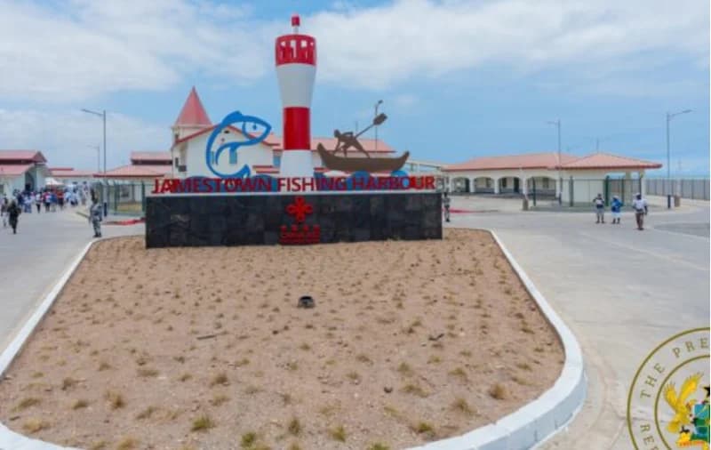 The image shows the entrance to the Jamestown Fishing Harbour in Accra, featuring a prominent monument with a red and white miniature lighthouse, a blue fish icon, and a silhouette of a fisherman. The facility appears modern and newly constructed, with several individuals visible walking around the grounds under a clear sky.