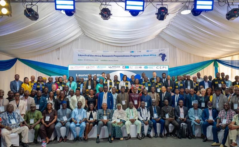 A large group of international delegates and health researchers pose for a group photo at the launch of the Africa Research Preparedness Program in West Africa. The event is held in a large hall in Accra, Ghana, decorated with blue and white ceremonial draping and professional stage lighting.