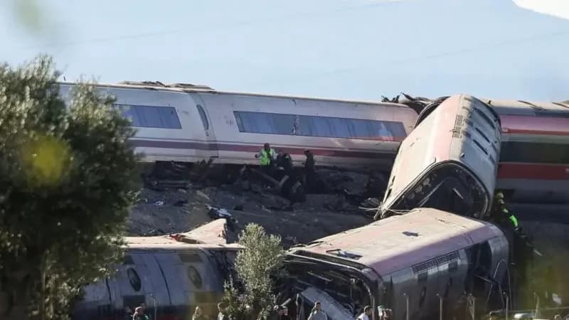 The image depicts the aftermath of a severe train derailment, showing multiple carriages crumpled and stacked against one another. Emergency personnel in high-visibility clothing are seen working near the wreckage, while onlookers gather at the site.
