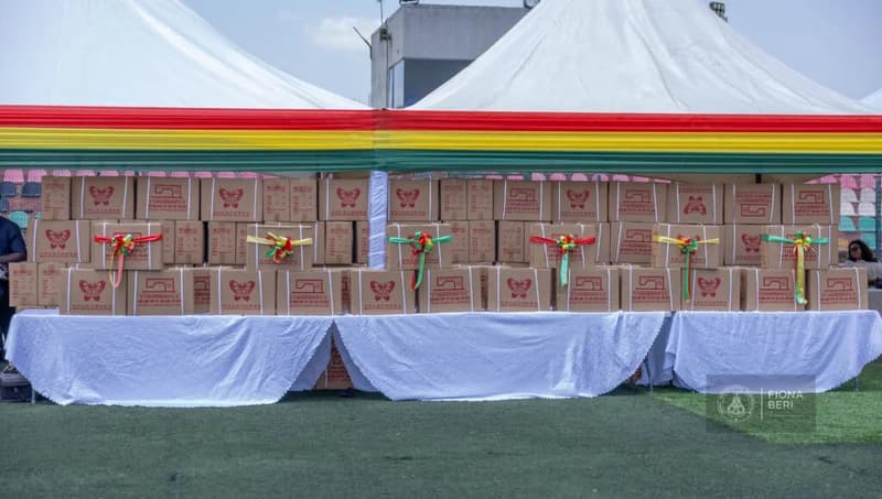 Numerous cardboard boxes containing sewing machines are stacked on tables covered with white linens under a large canopy. The event space is decorated with the Ghanaian national colors (red, gold, and green), and several boxes are adorned with decorative ribbons for a presentation ceremony.
