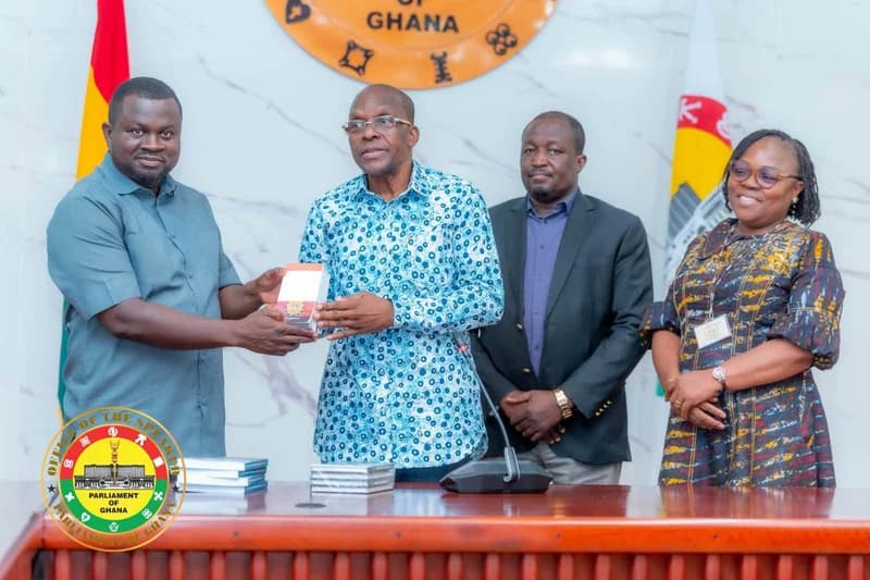 Speaker of Parliament Alban Bagbin is captured in a formal setting, receiving an item from a gentleman while two others look on. The group is positioned in front of the Parliament of Ghana seal and national flags, likely during a courtesy call or official presentation.