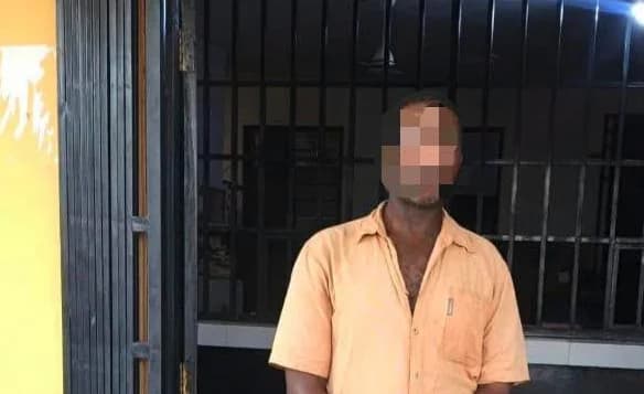 A man wearing an orange short-sleeved shirt stands in front of a black metal gate, likely at a police station. His face has been pixelated to protect his identity following his arrest.